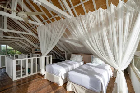 Bright loft bedroom with twin beds draped in sheer white canopy curtains, wooden beams, railing, and windows overlooking tropical greenery.